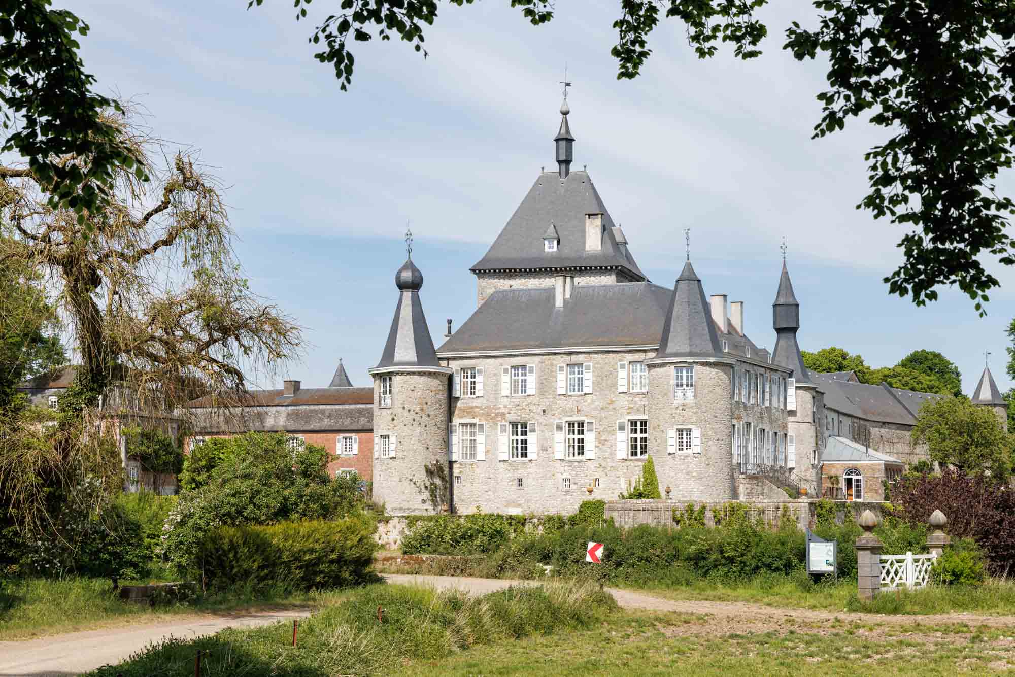 Château d’Hodoumont entouré de verdure, avec ses tours et façades en pierre sous un ciel clair.